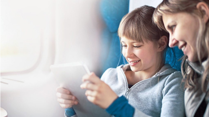 Eine Mutter sitzt mit ihrer jugendlichen Tochter im Zug. Sie schauen beide lächelnd auf ein Tablet. Das Licht scheint hell durch das Fenster und schafft eine ruhige, entspannte Atmosphäre.