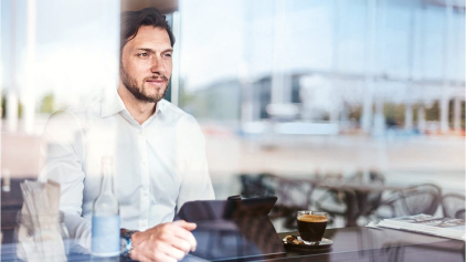 Ein Mann in den Dreißigern sitzt mit seinem Laptop vor einem Fenster in einem Café. Er sieht selbstbewusst, leicht lächelnd nach draußen. Vor ihm steht ein schwarzer Kaffee.