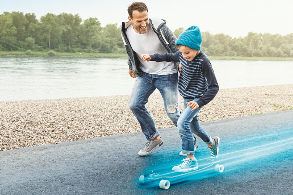 Vater rennt lachend neben seinem Sohn, welcher Skateboard fährt.