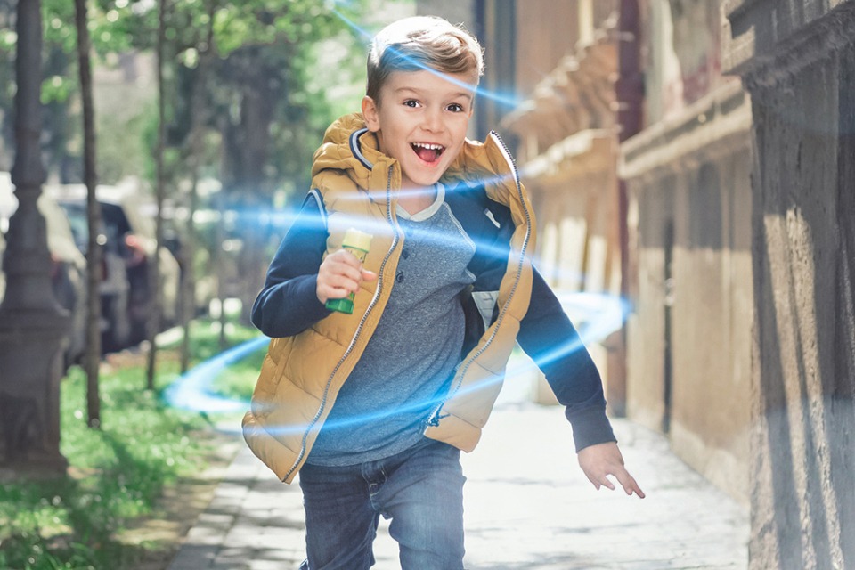 Ein kleiner Junge rennt auf dem Gehweg Richtung Kamera. Es ist ein sonniger Tag, er trägt eine Weste und freut sich.