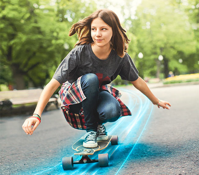 Ein Mädchen fährt in der Hocke Skateboard auf einem asphaltierten Weg in einem Park. Um sie herum sieht man Bäume, Büsche und eine Bank.