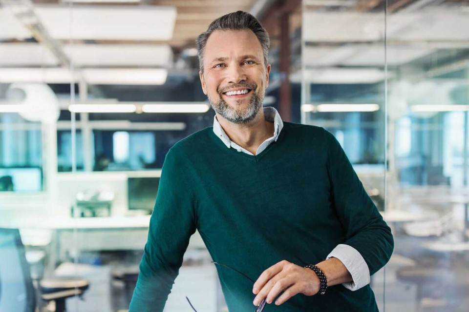 Mann in den Vierzigern im Business-Look mit Hemd und tannengrünem leichten Pullover steht im Büro und blickt offen lächelnd in die Kamera.