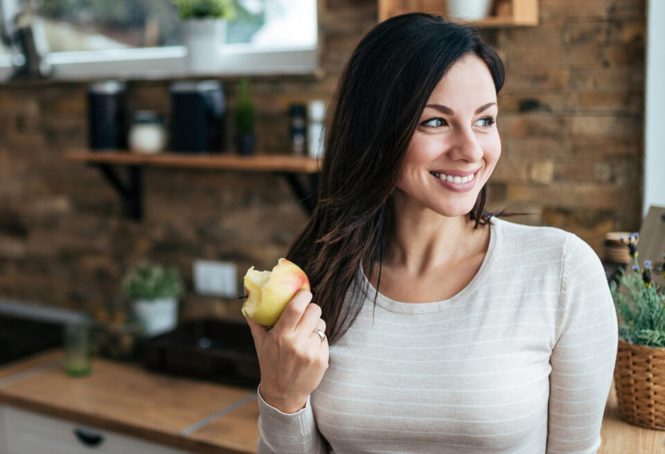 Eine junge Frau mit beige-weiß-gestreiftem Oberteil steht in der Küche, lächelt breit und hält einen Apfel in der Hand. 
