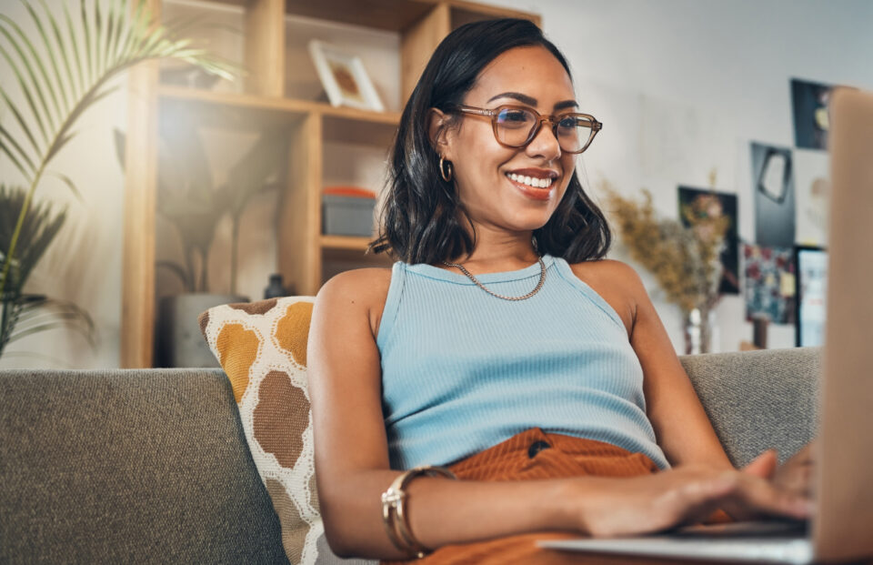 Eine junge Frau mit schulterlangem Bob und moderner Brille sitzt im hellblauem Tanktop und brauner Cordhose auf der Couch. Sie tippt lächelnd in ihren Laptop.