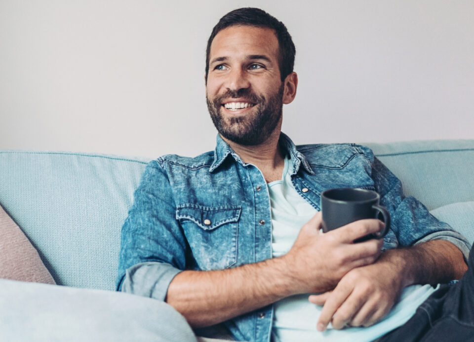 Ein junger Mann mit offenem Jeanshemd, hellblauem T-Shirt und Bart sitzt mit seiner Kaffeetasse entspannt auf der Couch. Er lächelt und genießt den Moment.