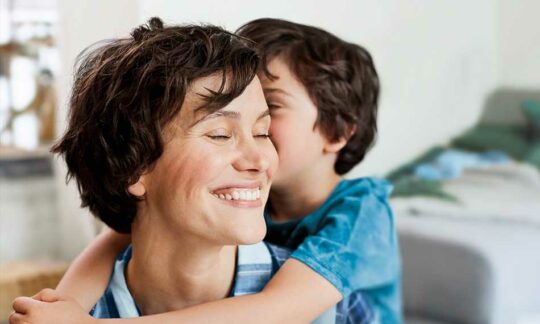 Junge Mutter wird von ihrem Sohn umarmt. Sie lächelt und zeigt ihre gesunden Zähne.