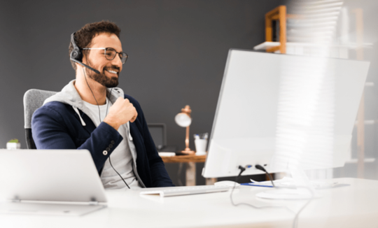 Junger Mann, der ein Headset trägt, sitzt zufrieden lächelnd in einem Online-Meeting vor seinem Bildschirm.