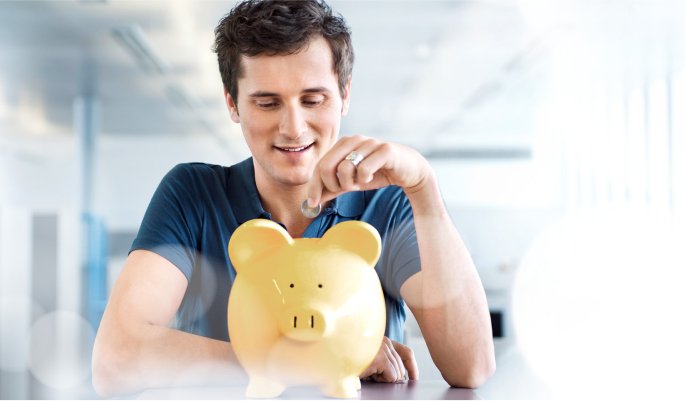 Ein junger Mann sitzt vor einem gelben Sparschwein und ist im Begriff, Geld einzuzahlen. Er lächelt entspannt.