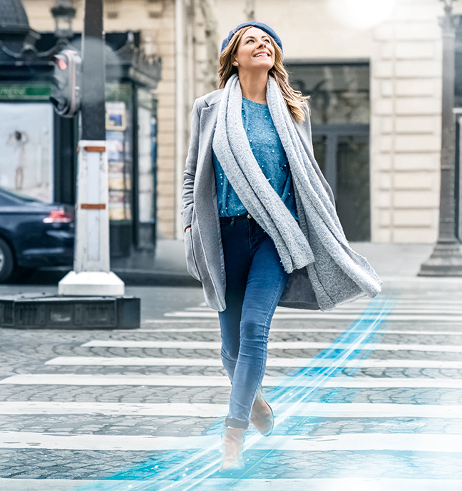 Eine Frau überquert lächelnd einen Fußgängerüberweg eine Straße. Sie schaut glücklich schräg nach oben, ihre Hände in ihrer graunen Manteltaschen.