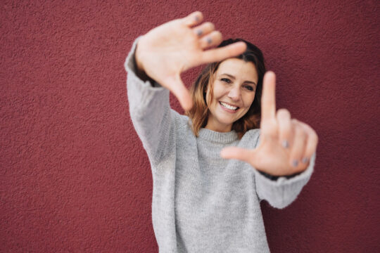 Frau mit grauen Pullover steht vor einer roten Wand und hält beide Hände in die Kamera und bildet damit einen Rahmen um ihr Gesicht.
