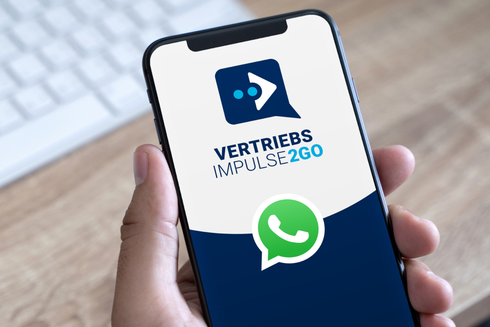 Eine Hand hält ein Smartphone, auf dessen Bildschirm das das Logo VertriebsImpulse2go und das WhatsApp-Logo zu sehen sind.